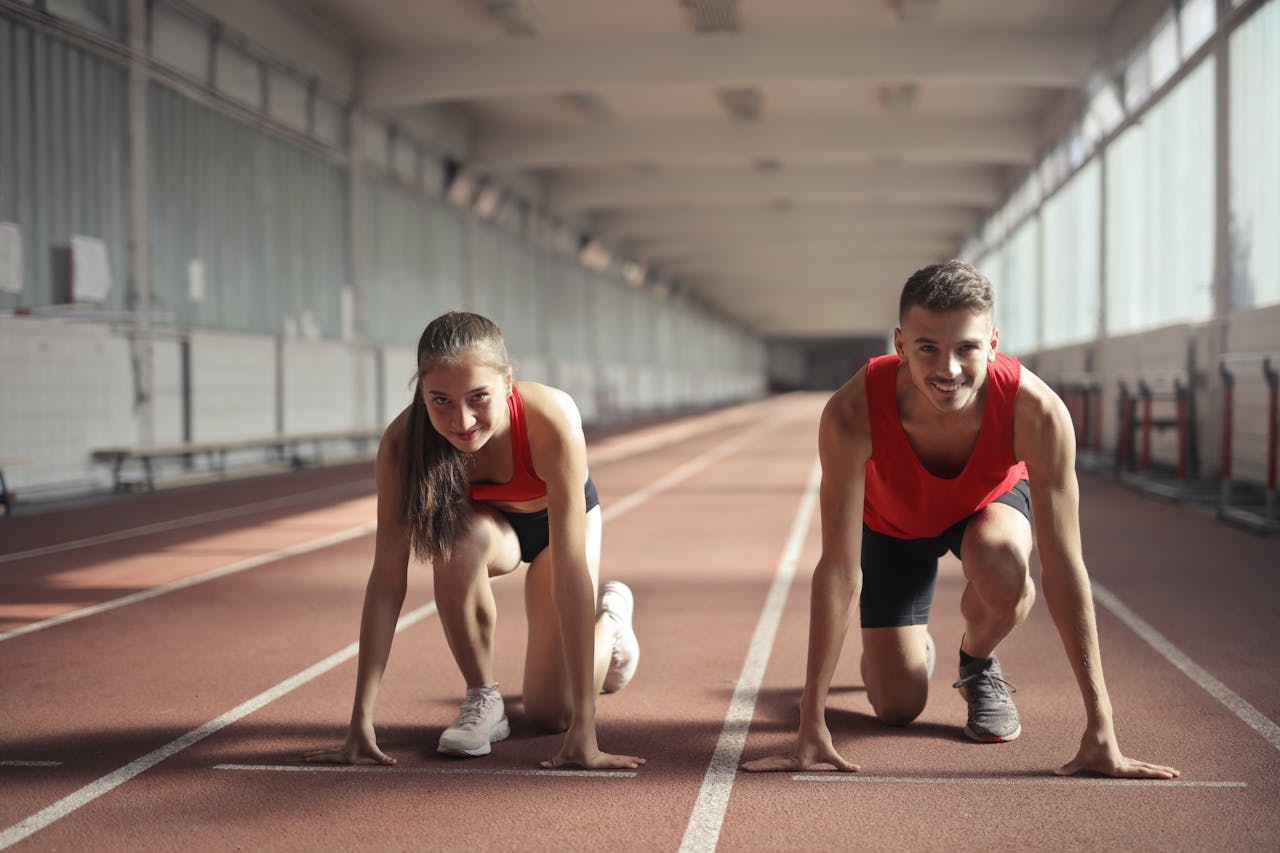 who-we-are Two athletes, a man and a woman, ready to sprint on an indoor track, embodying fitness and determination.