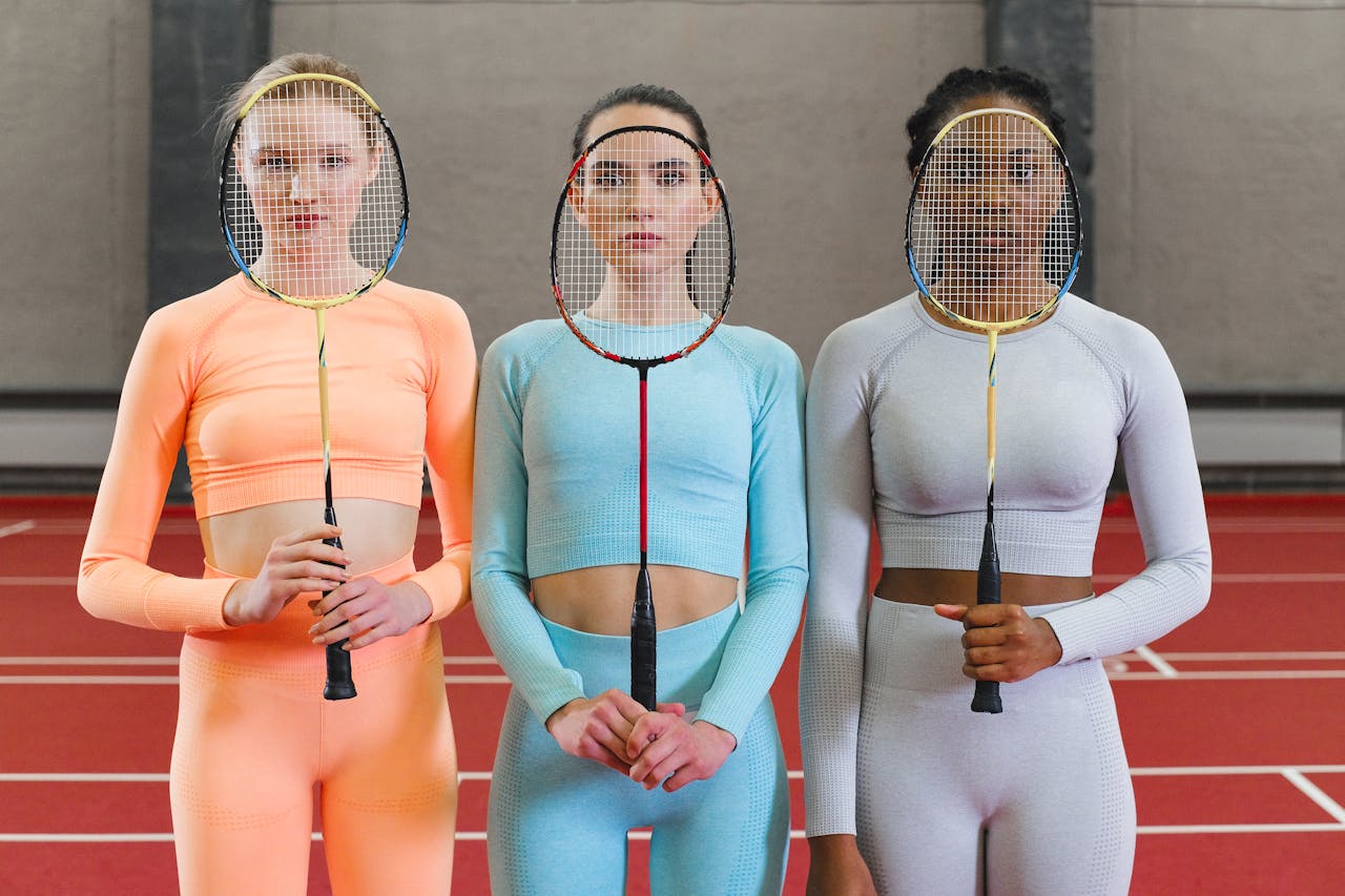 services-bg Three women athletes posing with badminton rackets in a gym setting, showcasing sports apparel and equipment.